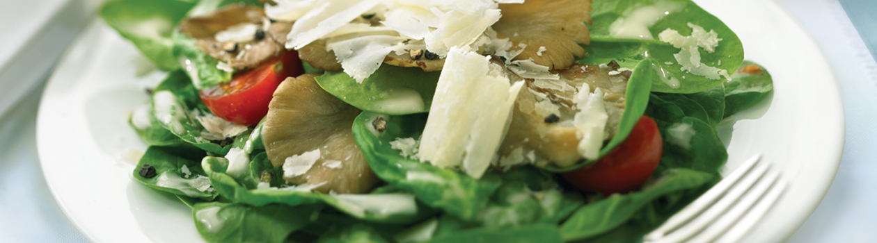 A close-up photograph of a bed of spinach topped with halved cherry tomatoes, grated parmesan cheese, small oyster mushrooms, and a drizzle of truffle and sherry vinaigrette.