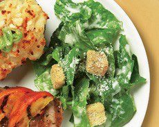 A close-up photograph of the corner of a ceasar salad dish containing grilled chicken, romaine lettuce, parmesan cheese, and croutons.