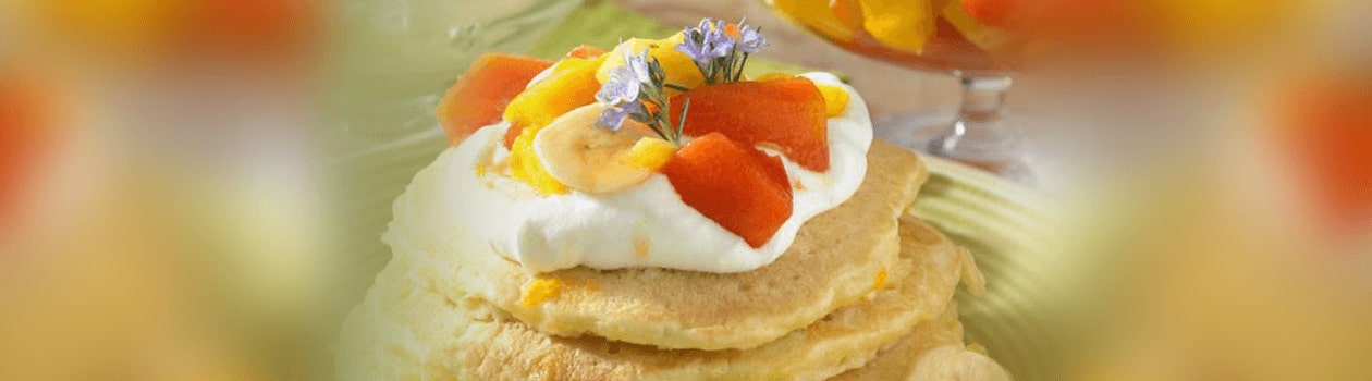 A close-up photograph of a stack of banana pancakes finishes with coconut yoghurt, banana slices, melon slices, and sweet lavendar sprigs.