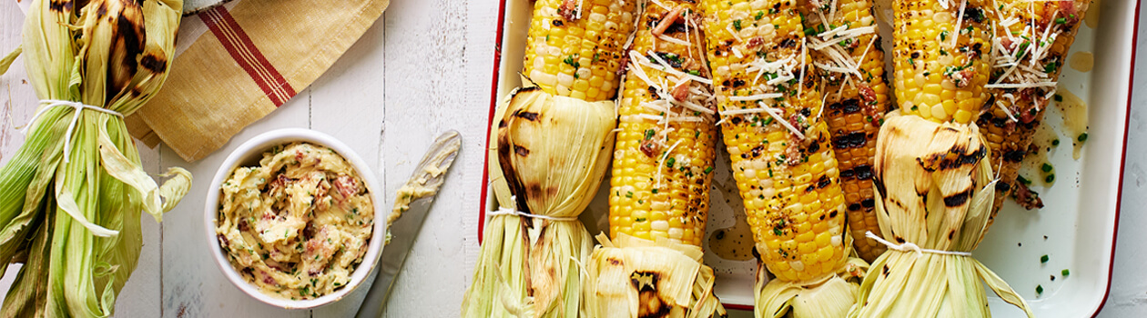 5 corn on the cob on a tray topped the all-dressed butter, a bowl filled with the butter sits to the left