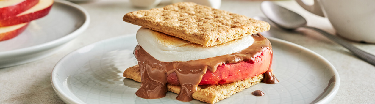 Apple, chocolate, and toasted marshmallow stacked between graham crackers on a plate. Apples, and marshmallows in the background.