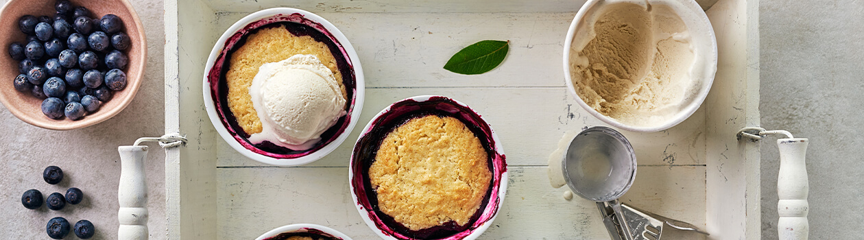 4 blueberry grants in ramekins on top of a wooden tray. Topped with a scoop of vanilla ice cream.