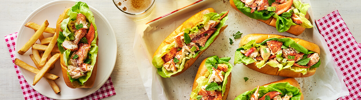 Five lobster rolls on a tray beside a ceramic plate holding a single lobster roll with a side of fries.